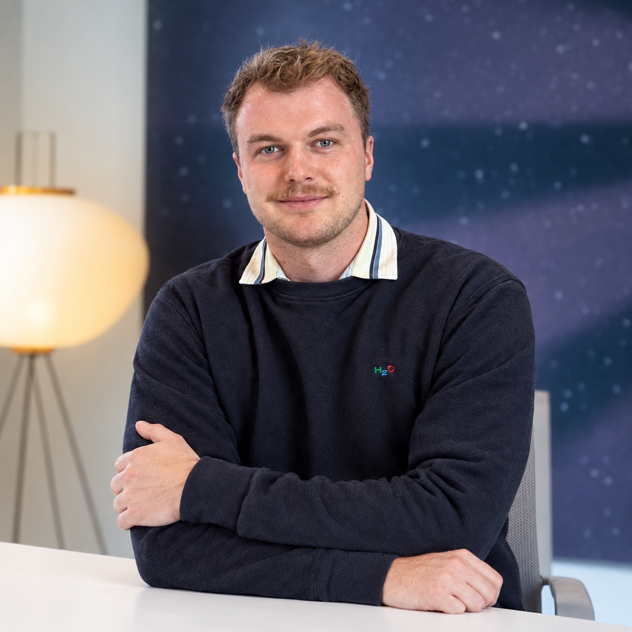 Man with crossed arms wearing dark sweater with H₂O logo, smiling at camera.